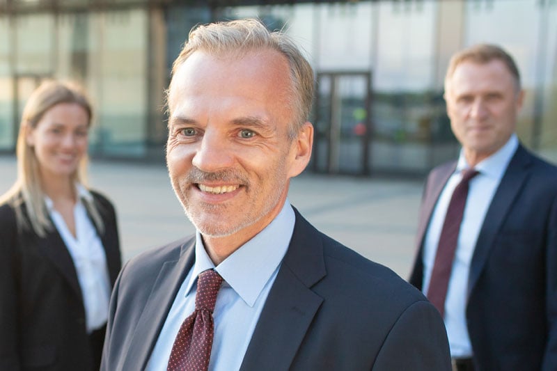 Portrait d’un entrepreneur souriant en costume – en équipe / Smiling entrepreneur in suit – with team in background